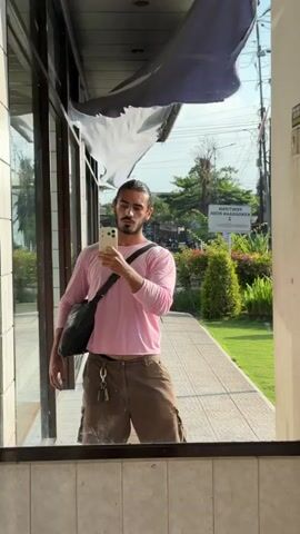 This is a selfie captured by a man standing in front of a mirrored surface, likely a window. He is holding a smartphone in his right hand, taking the photo. The man is wearing a long-sleeved pink t-shirt and brown cargo shorts. A black messenger bag is slung across his body. His hair is pulled back, and he has a beard.

The background visible in the reflection shows a sunny outdoor scene. There is a paved walkway, green grass, and lush vegetation including bushes and trees. A sign is visible in the distance with text in Indonesian, which appears to read "PENITIPAN KENDARAAN RODA DUA" which translates to "MOTORCYCLE PARKING". The sky is blue and clear, suggesting a warm, pleasant day. The overall mood appears casual and relaxed. The mirrored surface reflects the man and the outdoor environment, creating a layered composition. There are some signs of wear and tear on an overhead awning or fabric structure. The time of day appears to be daytime due to the bright sunlight.