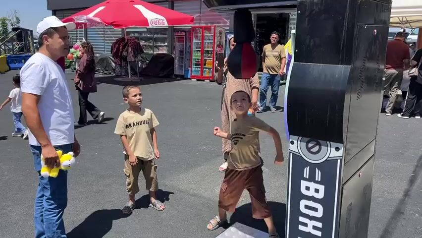 A group of people are gathered outdoors in what appears to be a market or fair. In the foreground, a young boy in a light brown t-shirt and khaki shorts is actively engaged with a boxing machine, his right fist raised as if to strike. To his left stands another young boy wearing a similar t-shirt and shorts, his arms outstretched as if in play or encouragement. A man in a white t-shirt and a white baseball cap stands to the left of the boys, holding a yellow stuffed toy in his left hand and looking towards the children. Behind the children and slightly to the right, a woman in a patterned dress is holding up a phone, seemingly recording the scene. A black boxing machine with the word "BOX" visible on its side is the central object of interaction. In the background, there are stalls with red awnings, one displaying a Coca-Cola logo. There are also several other people milling about in the background, and a vending machine filled with drinks. The ground is paved, and the lighting suggests a sunny day.