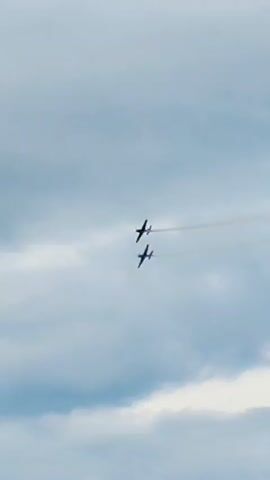 Two dark-colored propeller airplanes fly in formation against a cloudy blue sky. The planes are positioned one above and slightly behind the other. A faint white trail of smoke or vapor emanates from the lead airplane. In the lower right corner of the frame, a portion of a white, fluted column with a dark capital is visible, suggesting the footage was captured from the ground looking upwards. The scene appears to be outdoors, likely during daylight hours.
