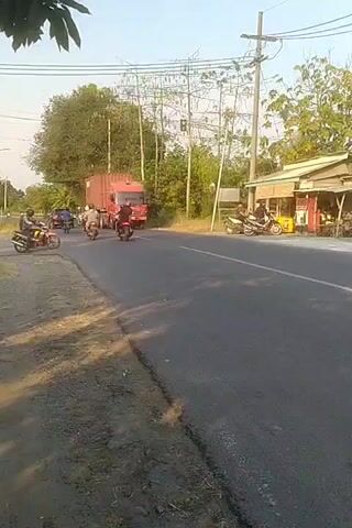 The video shows a red truck with a shipping container driving down a paved road. There are several motorcycles on the road, some moving with the truck and others in the opposite direction. To the right of the road, there is a small building with a roof and some people standing outside. There are trees and utility poles in the background. The sky is clear and appears to be daytime. The video is shot from a low angle, looking up at the truck and motorcycles.