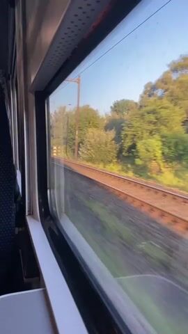 The video shows the view from a train window as it travels through a landscape with trees and power lines. The train appears to be moving at a moderate speed, as evidenced by the motion blur of the scenery outside the window. The interior of the train is visible on the left side of the frame, including a seat with a patterned fabric and a small table. The lighting suggests it is daytime, possibly late afternoon or early evening, with the sun casting a warm glow on the trees. The tracks are visible running parallel to the train.