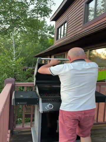 The video begins with a close-up of a man wearing a white polo shirt and red shorts standing on a wooden deck and closing the lid of a grill. The camera then pans to the left, revealing a scenic view of a lake and mountains at sunset. The sun is setting behind the mountains, casting a warm orange glow across the sky and the water. A small town with distinctive architecture is visible on the shore of the lake, with trees and foliage in the foreground. The overall impression is of a serene and picturesque evening.