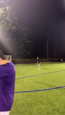 Two people are on an illuminated green artificial turf soccer field at night. One person in a purple shirt stands near a soccer goal, while the other person in a black shirt stands a short distance away and prepares to kick a soccer ball. The environment includes trees in the background and a blue sideline marking the field.
