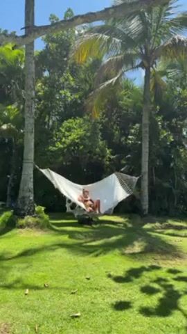 Here's a description of the video:

The video shows a woman relaxing in a white hammock. The hammock is suspended between two tall, slender trees in a grassy yard. Palm trees and other lush greenery surround the area. The sky is clear and blue. The woman is lying down, appearing relaxed and still.