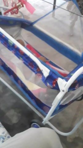 A baby lies in a blue and white crib, moving their hands to their face. The crib has a mosquito net. The floor is concrete.