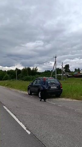 A person is standing next to a parked dark-colored SUV on the side of a paved road. The vehicle's trunk appears to be slightly ajar. The person is facing away from the camera and is dressed in dark pants and a blue jacket. The surrounding environment is rural, with green grass and trees visible. In the background, houses and utility poles can be seen under a cloudy sky. The camera is positioned slightly to the left of the road, capturing the scene from a moderate distance.