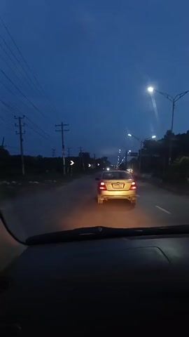 A video captures a view from inside a vehicle moving along a road during twilight. The sky is a deep blue, with visible power lines crisscrossing above. On the left, a utility pole with a sign is seen, followed by an expanse of green grass and distant trees. The right side of the frame shows the car's side mirror, reflecting the darkening sky and lights. Further down the road, headlights of oncoming vehicles and streetlights illuminate the path, indicating the presence of other traffic. The road surface is visible, with the car's dashboard in the foreground.