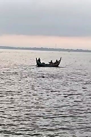 The video shows a small boat on a body of water. Three or four people are in the boat. The boat is propelled by oars. The background shows land and a cloudy sky. There are also a few other boats in the distance.