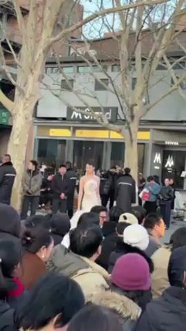 The video shows a crowd of people watching a fashion model. She is wearing a light-colored, one-shoulder dress with floral embellishments. She has dark hair pulled back. She walks confidently as people take photos with their phones. There are several men in black jackets standing around, likely security. The event is outdoors, with bare trees lining a paved area. Buildings with signs that say "M Grand" are visible in the background, along with a metal bridge.