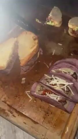A wooden board topped with sliced sandwiches and purple tacos is shown. A person picks up a sandwich and takes a bite. The person is a light-skinned young man wearing a black sweater that says “ALLSAINTS” in white lettering. He is facing the camera and makes a surprised expression after tasting the sandwich.