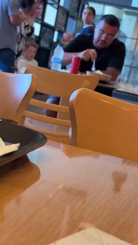 A medium shot captures a man in a black t-shirt and glasses at a table in what appears to be a restaurant. He is holding a fork and knife, actively eating. A red Coca-Cola can sits on the table in front of him. To his left, a younger boy in a light blue shirt is visible. In the background, several other people are present, including a woman in a floral dress and another person in a white shirt. The environment suggests a public dining space with wooden chairs and tables. The camera is positioned low, focusing on the table and the man, with the background slightly out of focus.