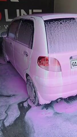 The video shows a pink car being washed at a car wash. The car is covered in pink foam. The car wash appears to be an open-air facility, possibly in Kokand City, Uzbekistan, given the context. The ground is wet and covered with foam, suggesting the washing process is in full swing.

Visible text includes "RED LINE CAR WASH" in English, and Russian text that translates to "FOAM ACTIVE CHEMISTRY." A license plate on the car reads "01 H819 TB". In the background, there are mops and buckets, as well as other cars and people, indicating a busy car wash. The lighting suggests it is daytime. The overall impression is of a routine car wash operation.