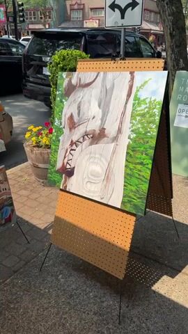 The video shows a collection of paintings displayed on easels along a sidewalk. In the foreground, a large painting of a totem pole with detailed carvings is visible, followed by a painting of Venetian architecture and canals, and another totem pole painting. Further to the right, a painting of a close-up of a weathered wooden face with dark markings is displayed. To the left, a painting of a branch with white flowers and green leaves is prominent. The paintings are displayed outdoors on a sunny day, with some people walking in the background and vehicles parked on the street. The environment appears to be a town or city with shops and buildings.