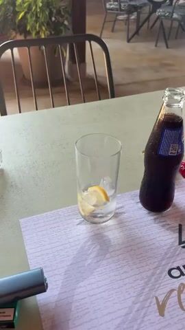 The video begins with a close-up of a glass with ice and lemon slices, a bottle of soda, and a red candy wrapper on a light-colored table. The camera then pans to the left, revealing a covered outdoor seating area with wooden beams and tables. In the background, several people are seated at tables, and some are standing and conversing. Further in the distance, a beach with umbrellas and people can be seen, and the ocean stretches to the horizon under a clear sky. The video also shows a woman in a bikini in the foreground, and a man in a white shirt and shorts seated at a table on the right, looking towards the beach.