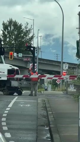 A person wearing an orange vest and a hard hat stands by a railway crossing gate. The gate is down, indicating that a train is expected or passing. The person appears to be overseeing the operation. A white truck is parked on the left side of the frame, next to the gate mechanism. Traffic signals are visible, with one showing a red light. An overpass and some trees are in the background. The ground is wet, suggesting recent rain.