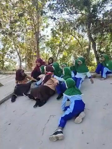 The video shows a group of young girls, dressed in school uniforms with headscarves, sitting in a semi-circle outdoors. They appear to be playing a game of tug-of-war, with some of the girls pulling on the arms of others. The environment is a sandy or dirt area with trees and foliage in the background. The girls are of various ages and seem to be enjoying themselves. One adult, likely a teacher, is also present, smiling and observing the activity. The lighting suggests it is daytime, possibly late morning or afternoon.