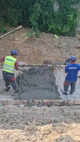 Two construction workers are pouring and spreading concrete into a formwork. One worker, wearing a yellow safety vest with "NDUCONST" printed on it, is using a shovel to move the concrete. The other worker, in a blue shirt with "NDUCONST" printed on it, is also using a shovel to spread the concrete. The scene appears to be a construction site, likely outdoors, with dirt and vegetation visible in the background. The lighting suggests it is daytime. The workers are focused on their task, indicating a professional work environment. The presence of "NDUCONST" on their clothing suggests they are part of the same company. The overall atmosphere is one of diligent labor.