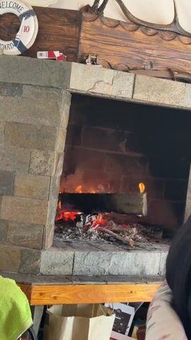 The video shows a stone fireplace with a small fire burning inside. There is a wooden shelf below the fireplace with various items on it, including a paper bag and some boxes. Above the fireplace, there is a wooden panel with a nautical-themed decoration and antlers. A person's head is visible on the right side of the frame. The audio includes speech.