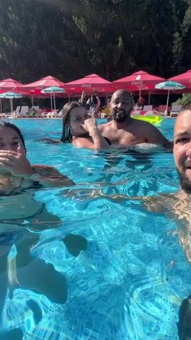 Four individuals are in a swimming pool. In the foreground, a woman in a camouflage bikini smiles and makes a peace sign as she looks toward the camera. To her right, a man in the water, partially submerged, looks at the camera. Behind them, a woman with long dark hair is being held by a man, also in the water. Both are looking towards the camera. In the background, the pool area has rows of red umbrellas and lounge chairs. The water is clear and blue, with sunlight reflecting on the surface. The bottom of the pool is tiled with a mosaic pattern.