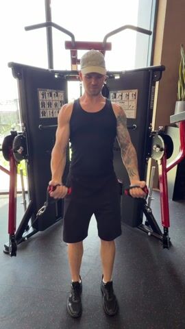 The video shows a man working out at a gym. He is wearing a black tank top, black shorts, and black shoes. He is using a multi-pulley machine to perform cable fly exercises for his chest. He has a light green cap and a tattoo on his left arm. The gym has large windows providing natural light.