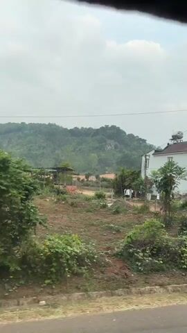 The video shows the view from a moving vehicle, likely a car or bus, traveling through a rural or suburban area. The landscape consists of houses, vegetation including trees and bushes, and a distant mountain range covered in trees. The sky is overcast. There is a utility pole near a dirt road. A few of the houses have fences and gates. A road marker is visible.