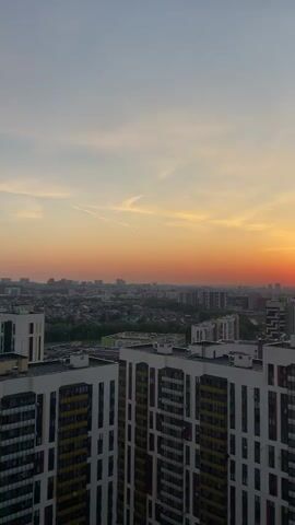 The video shows a sunset over a city. The sky is orange and red near the horizon, fading to a pale blue above. The sun is a bright red orb, just above the horizon line. Below the horizon, a cityscape stretches into the distance. There are many buildings, mostly modern apartment blocks, with some green spaces visible in between. The buildings in the foreground are predominantly white, with colorful accents like yellow, red, and black window frames. The camera is stationary, offering a wide shot of the scene.