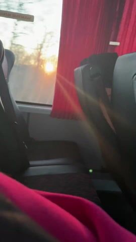 From the perspective of someone sitting on a bus, the camera looks towards a window. The sun, low on the horizon, shines brightly through the glass. A red curtain hangs alongside the window. Dark-colored seats line the bus interior. Part of a pink garment is visible in the foreground. A sign in a foreign language marks the emergency exit.