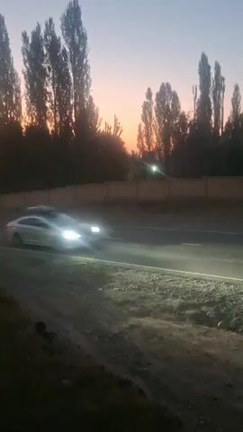 A silver car with a roof rack is driving on a paved road as the sun sets. The sky is a gradient of blue and orange, with tall trees silhouetted against it. A low wall runs along the side of the road.