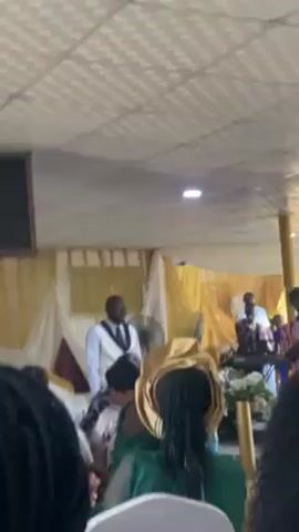 The video shows a man wearing a white suit jacket over a dark shirt and tie walking towards the camera. He is in what appears to be a church or event hall with people seated in the foreground. There is a stage with gold curtains and a keyboard player. The ceiling is white with a patterned design. The man walks in front of the seated crowd, then off to the right.
