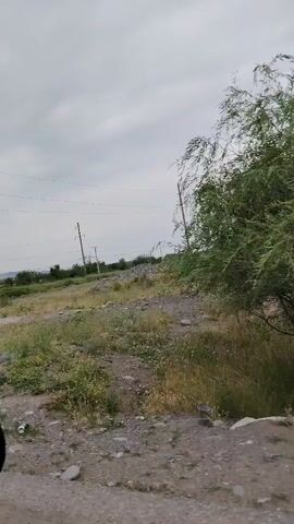 The video shows a person driving down a dirt road with a rearview mirror visible on the left side of the frame. The landscape is rural with fields, scattered trees, and a cloudy sky. There are power lines and utility poles throughout the scene, indicating the presence of electrical infrastructure. The ground is uneven and appears dry with patches of grass and gravel. The overall impression is one of a desolate, undeveloped area.