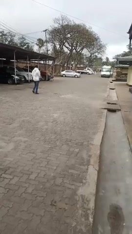 The video pans across a paved area, likely a parking lot or driveway, in front of a row of single-story buildings. The buildings appear to be low-rise with light-colored walls and darker trim, possibly offices or a residential complex. Some buildings have covered walkways or awnings extending from them. A drainage channel runs along the side of the paved area. In the background, trees and utility poles are visible. Several cars are parked in the lot, some under a covered structure, and a white van is seen further down the driveway. A person in a white hooded jacket is walking away from the camera towards the buildings on the left. A flag, appearing to be red, black, green, and blue, flies from a pole near one of the buildings. The sky is overcast.