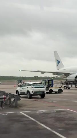 A cargo airplane with the logo "BLUE DART" on its side is parked on an airport tarmac under a cloudy sky. Several ground vehicles are near the plane, including a white electric car, a small tractor-like vehicle, and a cart with a covered cargo area. A few individuals wearing high-visibility vests are visible, some appearing to be working near the aircraft. The airplane has its landing gear deployed.