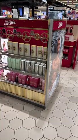 The video pans across a well-lit duty-free shop, likely an airport or a large department store, displaying a Jean Paul Gaultier perfume counter. The counter is elaborate, with a rich red backdrop adorned with golden gears, evoking a steampunk or industrial aesthetic.

The shelves are meticulously arranged with various Jean Paul Gaultier perfumes and gift sets. On the top shelf, large cylindrical containers with a sunburst design are visible. Below them, rows of pink and mint green packaging, along with silver cylindrical containers, showcase different fragrance lines. The lower shelves feature smaller perfume bottles and more pink gift boxes, some labeled "SCANDAL." The overall impression is one of luxury and sophistication.

The floor is tiled with hexagonal patterns in light and dark grey. In the background, other retail displays can be seen, including a prominent pink stand labeled "KYLIE COSMETICS," featuring makeup products and a director's chair. Further back, a large screen displays an advertisement, and a sign for "CHANEL" is visible. The presence of other brand displays and general store layout suggest a busy retail environment.

Although no specific activity is clearly depicted beyond the display of merchandise, the setting implies a retail experience where customers can browse and purchase perfumes and cosmetics. The lighting is bright and consistent, suggesting it is daytime, and the indoor setting means weather is not a factor. The overall mood is commercial and aspirational, focusing on the allure of high-end fragrance brands. The visible text includes "Jean Paul Gaultier," "SCANDAL," "KYLIE COSMETICS," and "CHANEL," reinforcing the brand-focused nature of the scene.