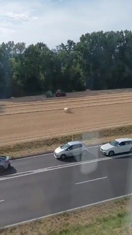 A tractor is working in a field of dry crops, possibly harvesting or baling hay. A single round bale of hay is visible in the field. Several cars are driving on a road that runs parallel to the field. The scene is set against a backdrop of dense green trees under a partially cloudy blue sky.