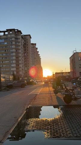 The video shows a street scene at sunset, with tall apartment buildings lining the street. Cars are parked along the road. The sun is low on the horizon, casting long shadows. In the foreground, a puddle reflects the sunlight. A person is standing by an electric scooter. The sky is clear and blue.