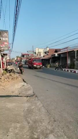 A red dump truck drives down a paved road. The truck is on the right side of the road. There is a sidewalk to the left of the road, with rocks and debris on it. A person on a motorcycle rides on the sidewalk, to the left of the truck. Buildings line the right side of the road. The sky is clear and blue.