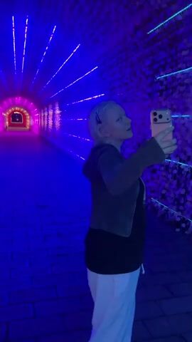 A woman stands in a tunnel taking a selfie. The tunnel has a brick floor, and the walls and ceiling are adorned with white flowers and illuminated by a rainbow of light that changes color throughout the video. The woman is blond, wearing a black t-shirt, a dark gray jacket, and light-colored pants. She holds an orange cell phone up to take the picture.