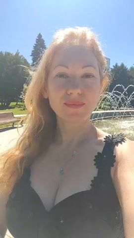 The video is a close-up selfie of a blonde woman smiling at the camera. She is wearing a black dress with a plunging neckline and intricate floral detailing on the straps. A light blue pendant necklace is visible around her neck. Her hair is long and wavy, falling over her right shoulder. The background appears to be a park on a sunny day. A fountain with water jets is visible to her right, and trees and a bench can be seen in the distance. The sky is clear and blue.