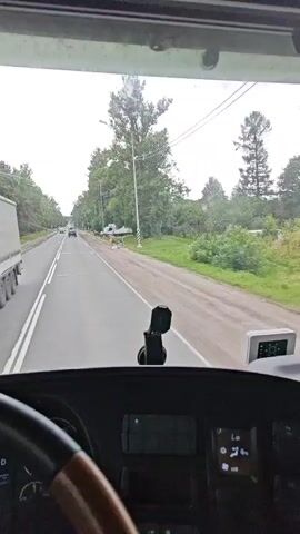 A truck driver's POV video shows a road with trees on either side. The truck is traveling on the right side of the road. On the right side of the road, a white truck with its rear door open appears to have crashed into a utility pole. Plants and debris are scattered around the crashed truck.