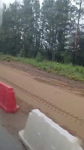 The video shows a view from a moving vehicle. The vehicle appears to be on a paved road, with a construction barrier on the left side. Beyond the barrier, there is a dirt road with tire tracks. The dirt road is lined with trees. The sky is overcast.