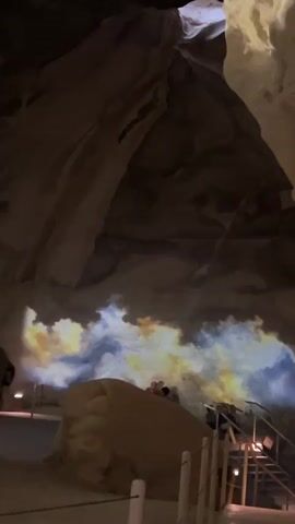The video pans upward in a dimly lit cavern. On the lower portion of the cave wall, a projection displays swirling colors of blue, yellow, and white, resembling clouds or smoke. In front of the projection, a large, light brown rock formation is visible. Several people are seated on or near this rock formation, their backs to the camera, and appear to be watching the projection. To the right of the rock formation, a set of stairs with railings leads further into the cave. The cave ceiling is rugged and appears to be lit from above by a bright opening.