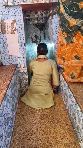 A woman in a light green salwar kameez kneels and prays facing a small alcove inside what appears to be a temple.  The alcove is tiled in blue and white mosaic tiles and has a trishul (a trident symbol associated with the Hindu god Shiva) on the back wall. Light shines brightly from within the alcove. The woman is kneeling on a beige tiled floor. Another woman wearing a yellow and green sari stands nearby on the right side of the frame.  The camera angle is at eye-level from behind the kneeling woman. The space is small and enclosed. The surrounding walls are also tiled with a similar mosaic pattern.  Sounds of children's voices can be heard in the background.