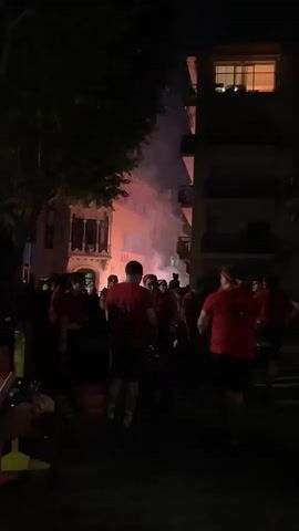 A crowd of people wearing red shirts and shorts are gathered on a street at night. Smoke or fog is visible in the air, illuminated by pink and orange lights from the surrounding buildings. Some people are holding their phones up, possibly recording the event. The atmosphere appears festive or celebratory. The buildings in the background are illuminated, and one building on the right has visible windows with lights on. Trees are visible on the left side of the frame.