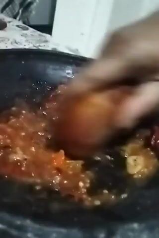 A hand is mashing tomatoes and chili peppers in a stone mortar. The tomatoes are red and chunky, and the peppers are small and red. The mixture is a vibrant red color and appears to be slightly watery. The mortar is dark gray and appears to be made of stone. The background is blurry, but it looks like a kitchen counter with some white objects. The lighting is dim and natural.