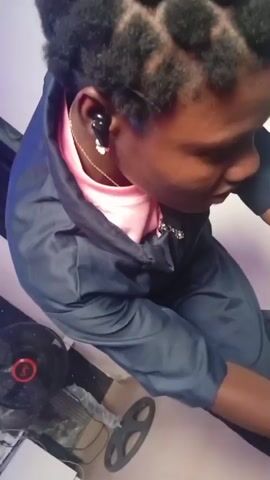 The video is a close-up, high-angle shot of a person with dark skin and braided hair, wearing a pink shirt and a dark blue jumpsuit. The person is looking down and appears to be engaged in an activity. There is a black earbud in their left ear and a silver earring. In the lower left corner of the frame, a black fan with a red circular element is visible, along with what appears to be a black object with a circular opening. The lighting is dim, with a light-colored wall in the background. The video is characterized by its shaky camera movement and lack of clear focus.