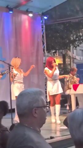 A live outdoor performance is shown. Two women in white minidresses and white boots are singing and dancing on a stage. One woman has blonde hair and the other has red hair. A man wearing a gold jacket is playing a white keyboard behind them. The stage has a white curtain in the background with red and blue lights shining on it. The performance is taking place at night, with trees and buildings visible in the background. A few audience members are visible in the foreground.