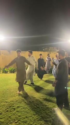 A group of men are dancing outdoors at night on a grassy lawn. The men are wearing traditional clothing, which appears to be shalwar kameez. Some of the men are dancing in a circle while others are watching. There are lights illuminating the area, and a wall in the background. One man in the foreground is recording the event with his phone. The overall atmosphere appears to be festive.