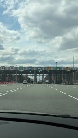 The video, filmed from inside a vehicle, captures the approach to a multi-lane toll plaza in Bizyar, Russia. The sky above is partly cloudy, indicating daytime conditions.

The toll plaza features numerous lanes, with overhead gantries displaying signs for payment methods and lane status. Several lanes are marked with green arrows, indicating they are open, while others show red 'X's, signifying closure. Prominently displayed Cyrillic text above the lanes identifies payment options: blue signs with a transponder icon read 'Т-РАНСПОНДЕР' (Transponder); orange signs with a card icon indicate 'КАРТА' (Card); and yellow signs with coin/cash icons denote 'НАЛ.' (Cash).

The road leading to the plaza is asphalt with clear white lane markings. Orange and white traffic cones line the left side of the approach. A white car is visible in an adjacent lane to the left, also approaching the booths. Beyond the toll structure, a treeline or forest is visible in the distance, suggesting a somewhat rural or suburban setting.

The car from which the video is shot slowly proceeds towards and then through one of the open toll lanes, smoothly navigating the payment point. No specific individuals are interacting at the booths, suggesting automated or fast-pass systems are in use for the visible lanes. The overall atmosphere is calm and functional.