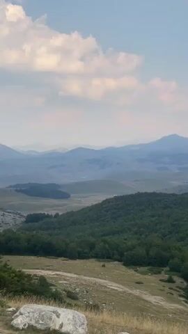 This is a vertical video that pans across a mountainous landscape. In the foreground, there is a grassy slope with scattered rocks and some dry grass. A dense forest of green trees covers the middle ground, with the trees appearing to be deciduous. Beyond the forest, rolling hills and mountains stretch into the distance, with the furthest peaks appearing hazy and blue. The sky is partly cloudy with some fluffy white clouds against a light blue background. The overall impression is one of a vast, natural landscape, likely captured during daytime. There are no people or animals visible in the video.