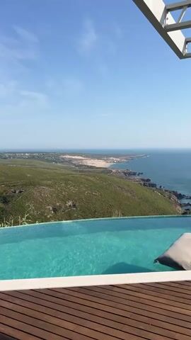A drone shot flies over a beautiful infinity pool with a pillow floating in it. The pool is next to a wooden deck and overlooks a scenic coastline with green hills and the blue ocean.